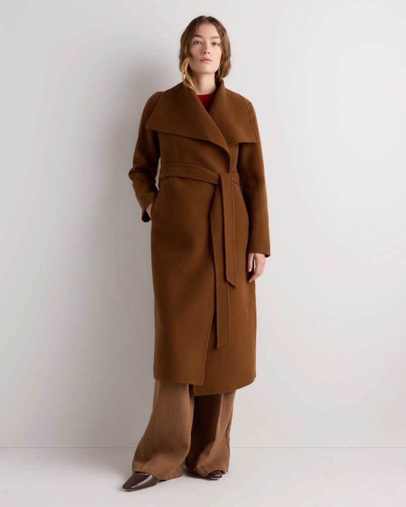 A woman wearing a brown wrap coat with a wide collar and a tie waist, paired with beige wide-leg trousers and brown shoes, standing against a neutral background.