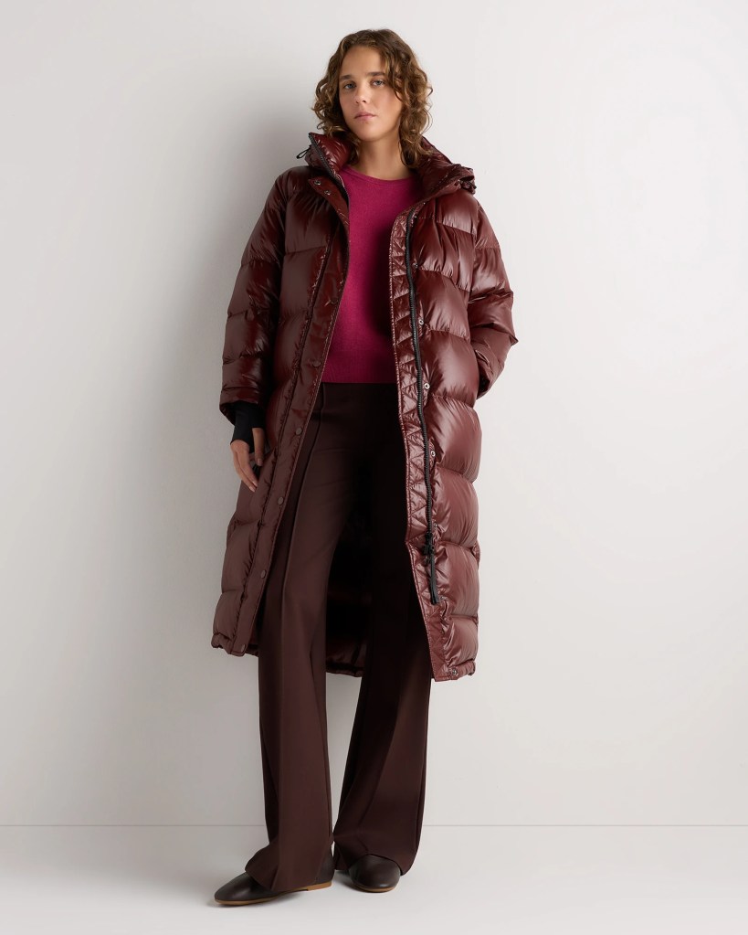 A woman wearing a long, burgundy puffer coat, paired with a pink sweater and brown wide-leg pants, standing against a light background.