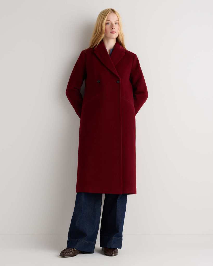 A model wearing a long burgundy coat with a double-breasted design, standing with hands in pockets against a light background.
