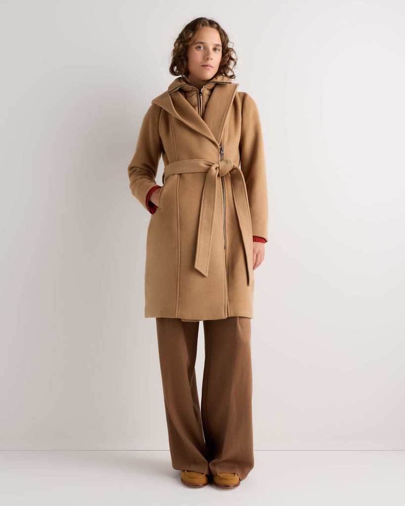 A young woman wearing a tan belted coat over a brown outfit, posing against a neutral backdrop.