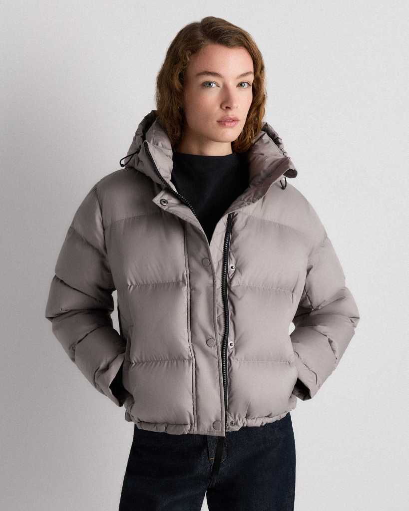 A woman wearing a light gray puffer jacket with a hood, posing against a neutral background.