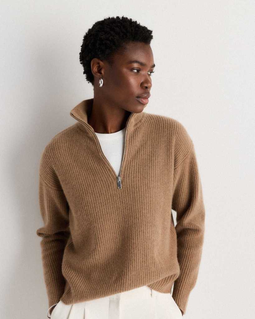 A young woman with short, curly hair is wearing a cozy brown ribbed zip-up sweater, looking thoughtfully to the side against a neutral background.