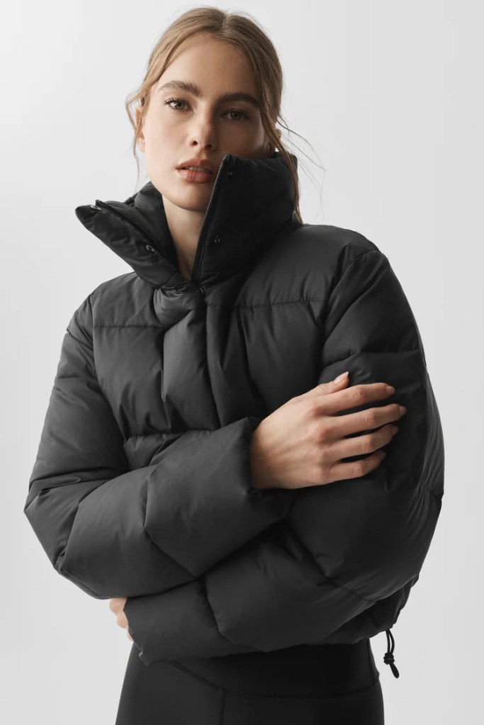A woman wearing a black puffer jacket with a high collar, posing against a neutral background.